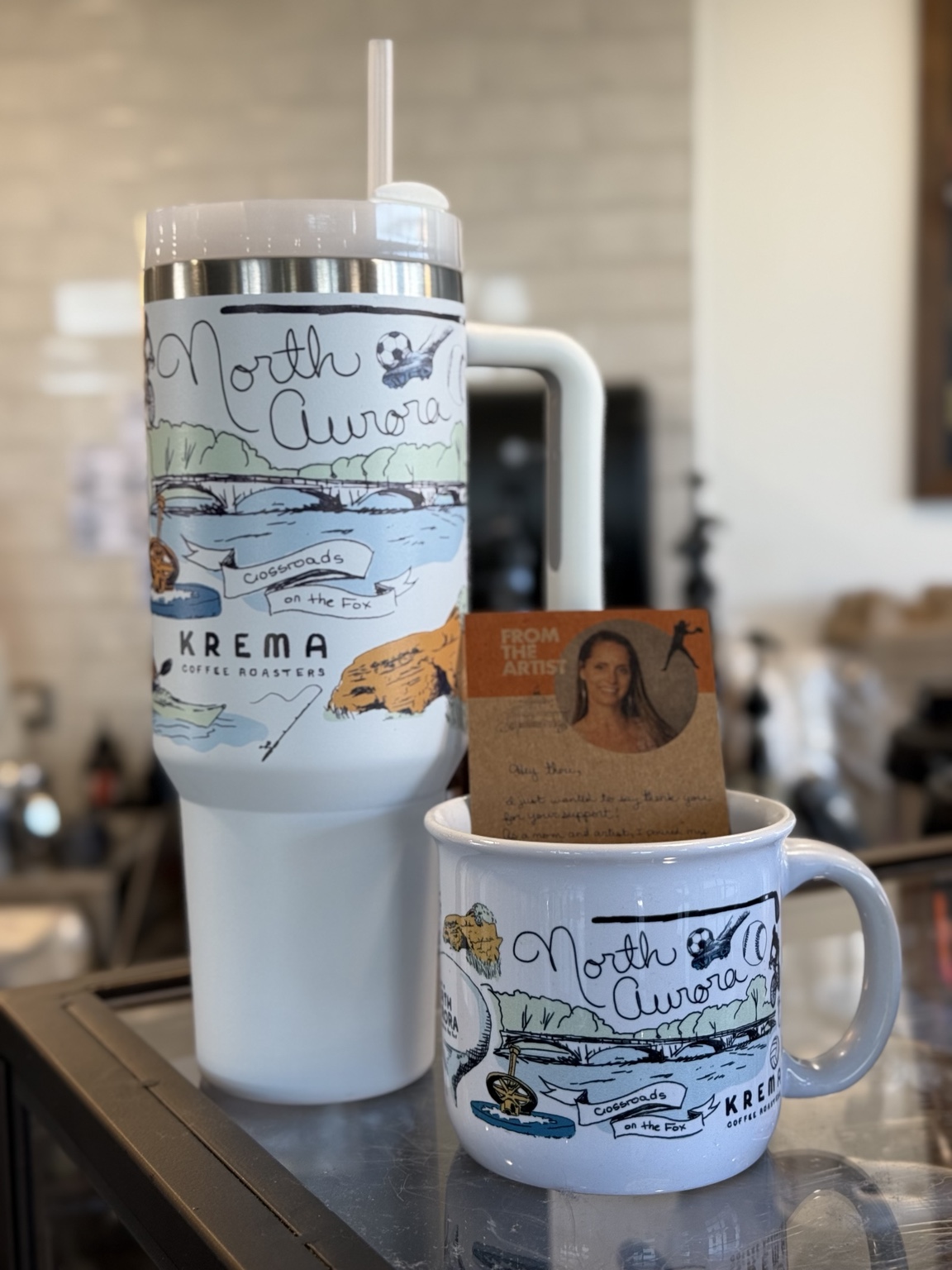 The hand-drawn North Aurora mug and matching travel tumbler displayed at Krema Coffee